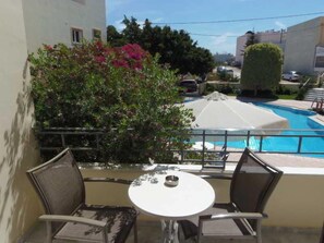 Un balcón con mesa y sillas que da a una piscina, rodeado de vegetación y edificios.