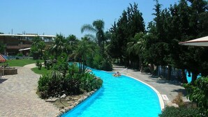 A swimming pool with a curved edge, surrounded by lush greenery and a paved walkway. - Atlantica Yakinthos Hotel (Chania)