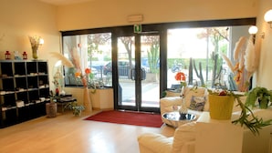 A reception area with a white sofa, a black shelving unit, and a red carpet leading to the entrance. - Hotel Provenza (Ventimiglia)