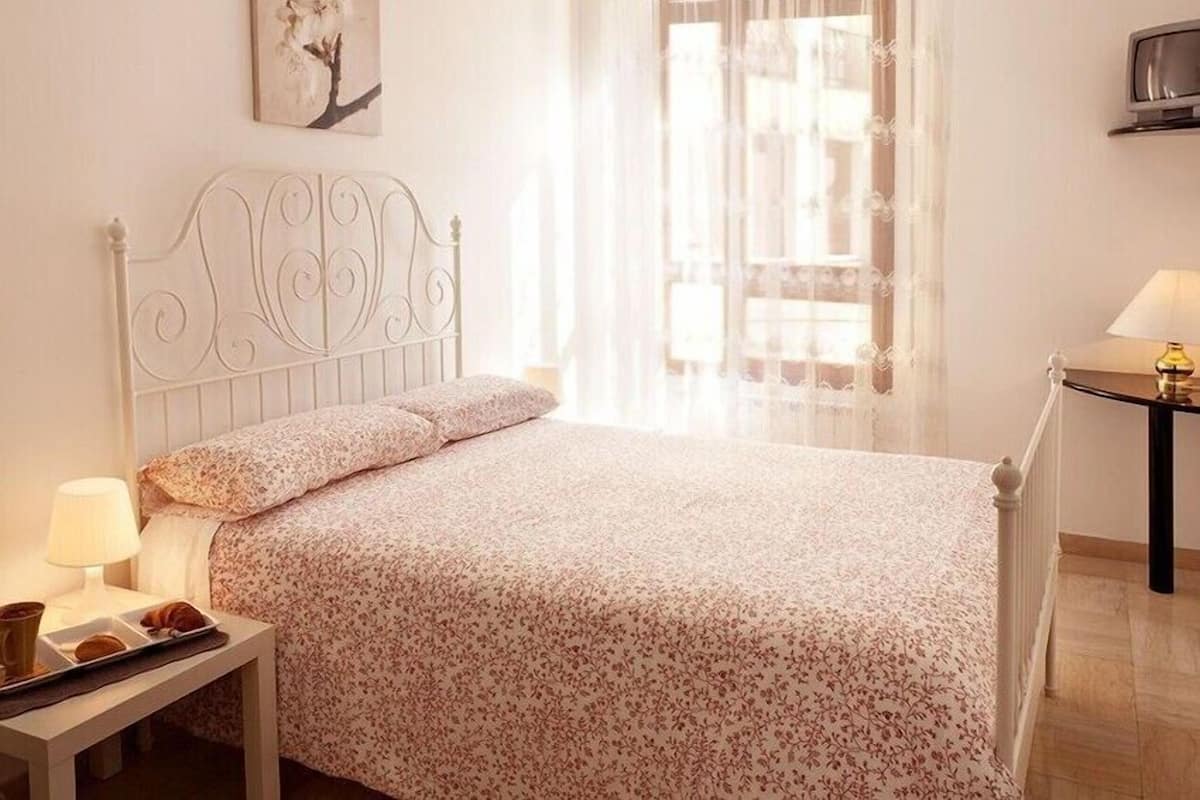 A bedroom with a bed, a bedside table with a lamp, a small table with a cup and a plate, and a window with sheer curtains.