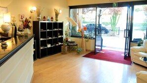 A reception area with a counter, shelves with decorative items, a seating area with a red rug, and a view of the outside through a glass door. - Hotel Provenza (Ventimiglia)