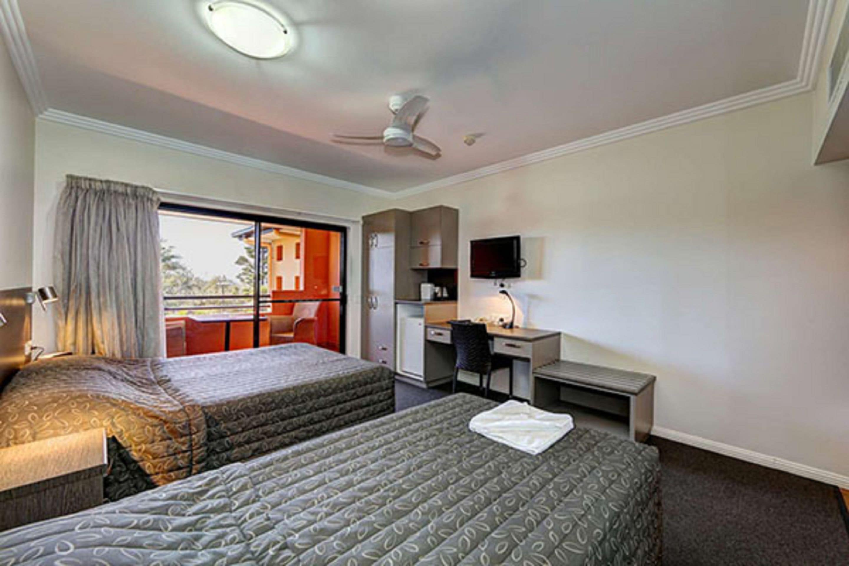 A hotel room with two beds, a desk, a chair, a television, and a ceiling fan.