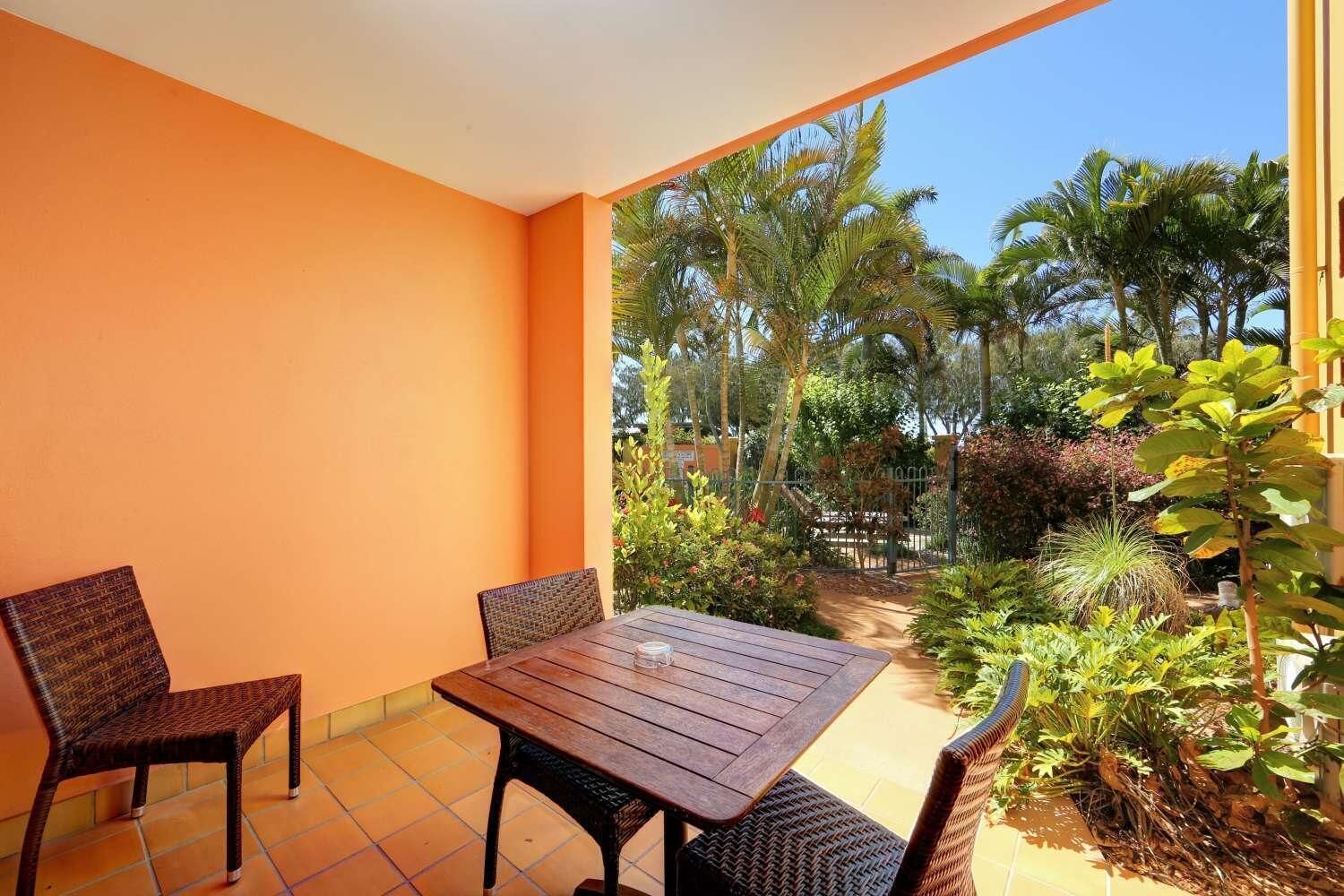 Un balcón con mesa de madera y sillas de mimbre, rodeado de paredes anaranjadas y vistas a plantas tropicales.