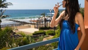 Una mujer con vestido azul parada en un balcón con vista a una playa con palmeras y una valla de madera.