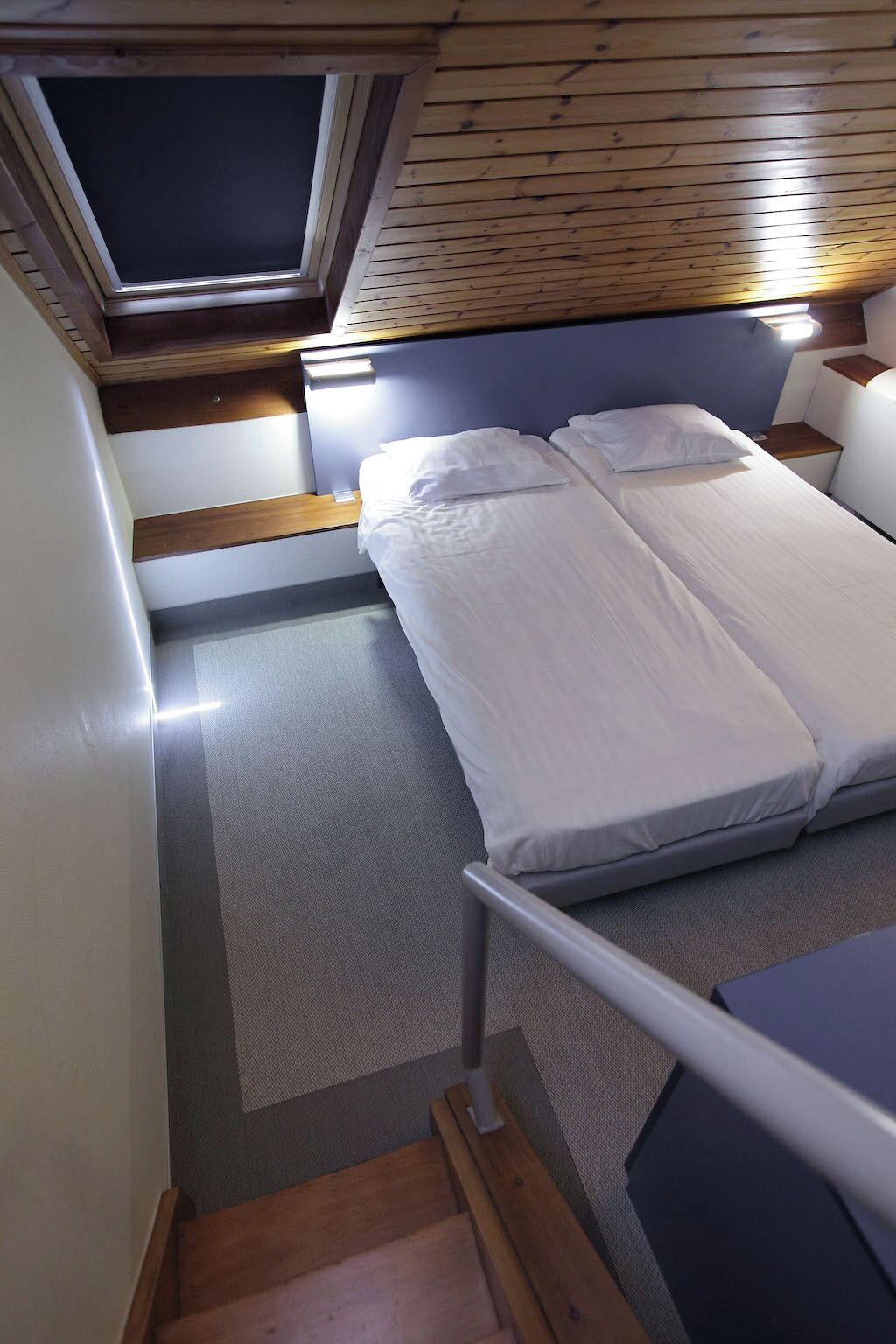 A compact room with a bed, a small table, a window, and a wooden ceiling.