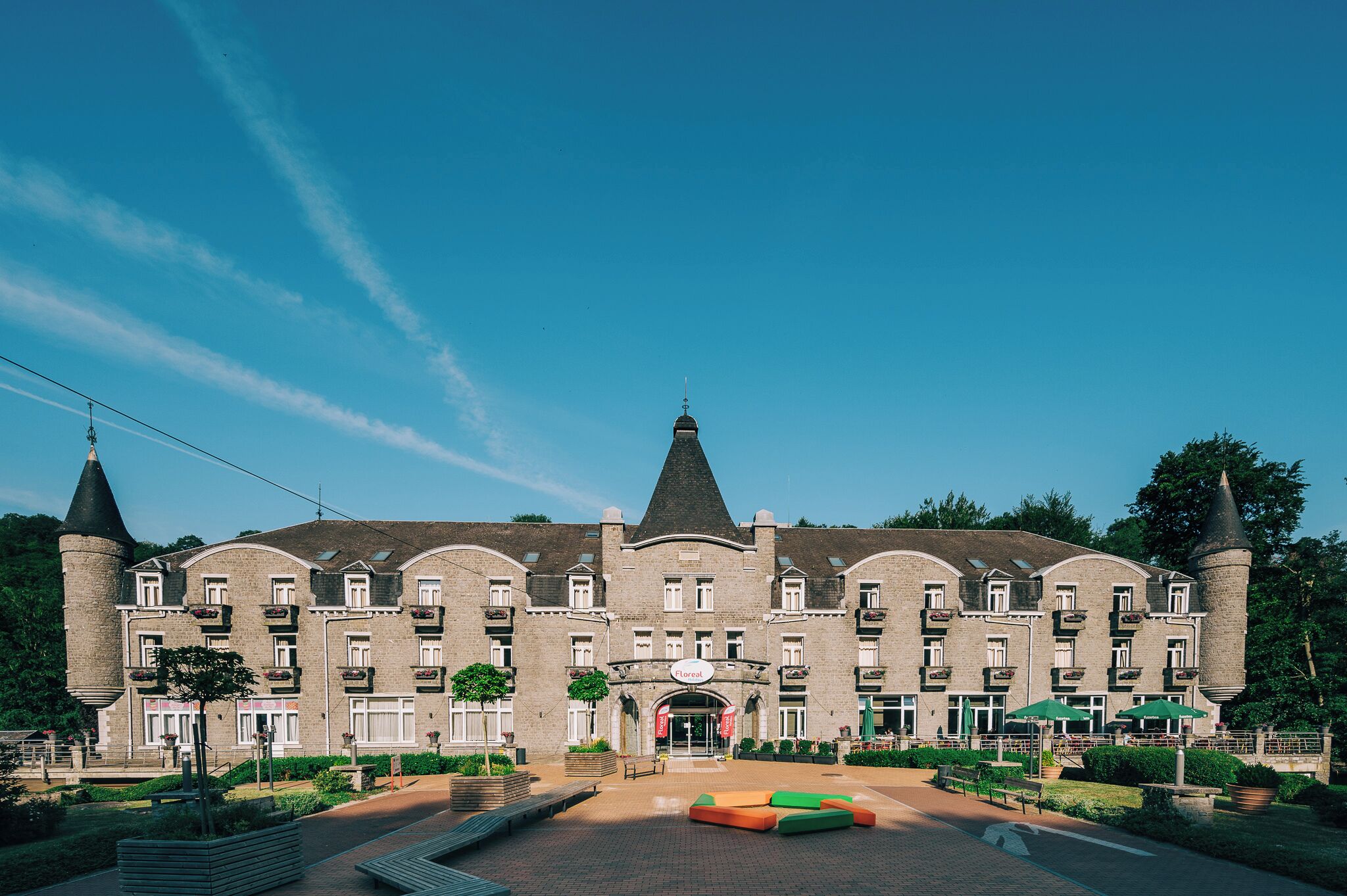 A large, historic building with a central tower, multiple balconies, and a paved courtyard with seating areas.