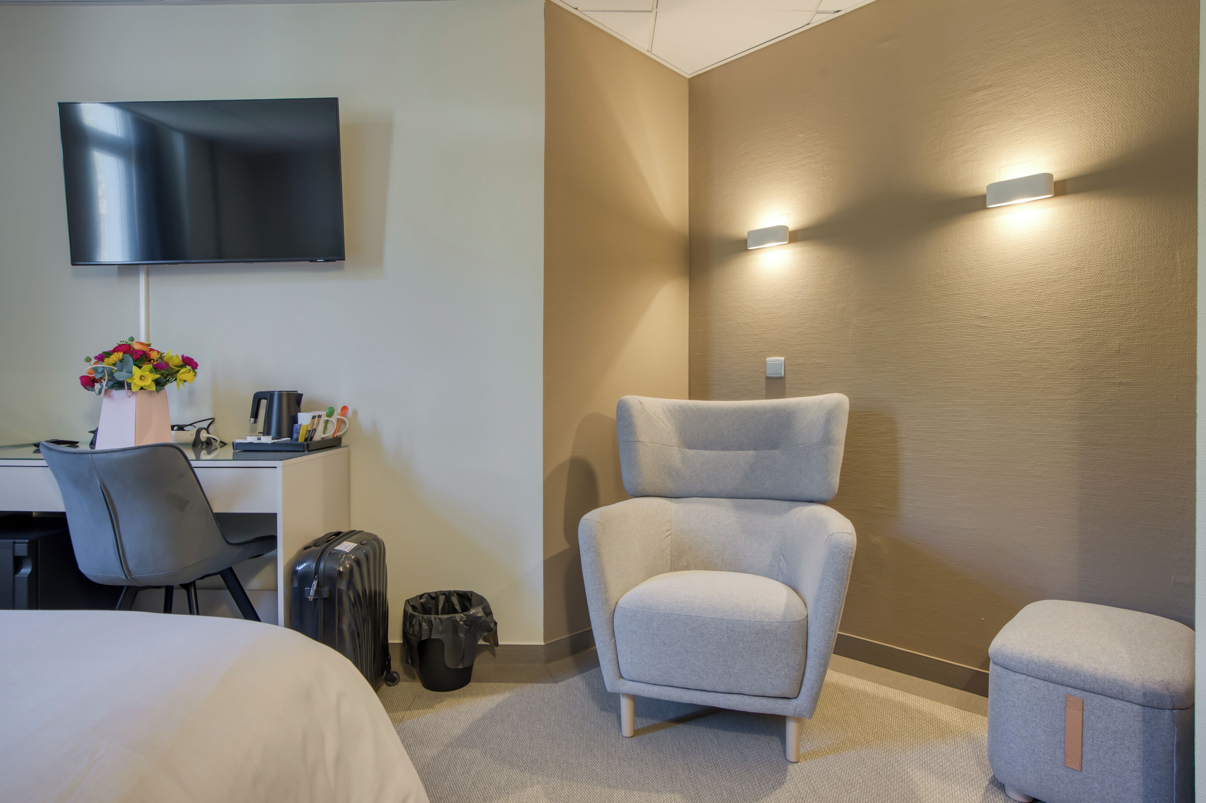Ett hotellrum med en säng, ett skrivbord med en tv, en grå fåtölj, en liten pall och en papperskorg.