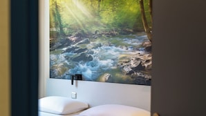 A bathroom with a wall-mounted sink, two white pillows, and a painting of a forest stream.