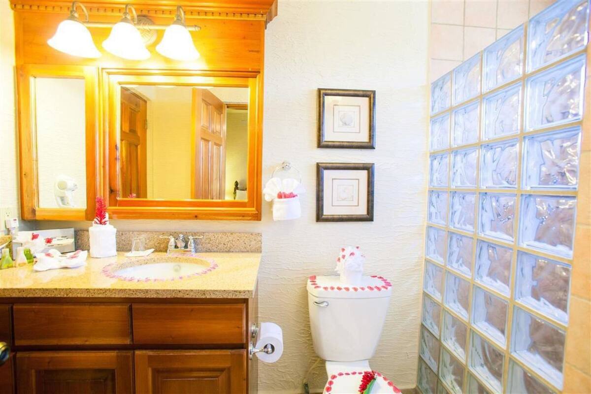 A bathroom with a wooden vanity, a large mirror, a toilet with a decorative cover, and a tiled wall with framed pictures.