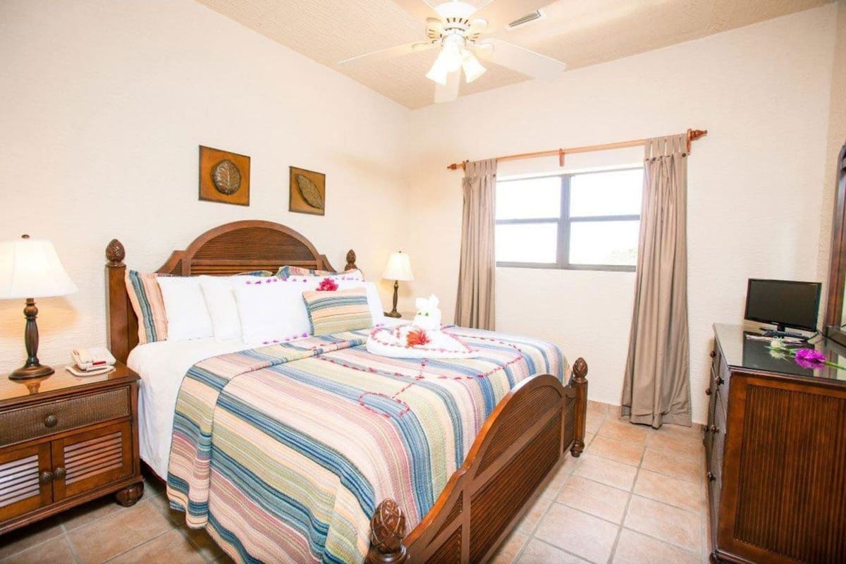 A bedroom with a bed, bedside tables, a desk with a computer, a ceiling fan, and a window with curtains.