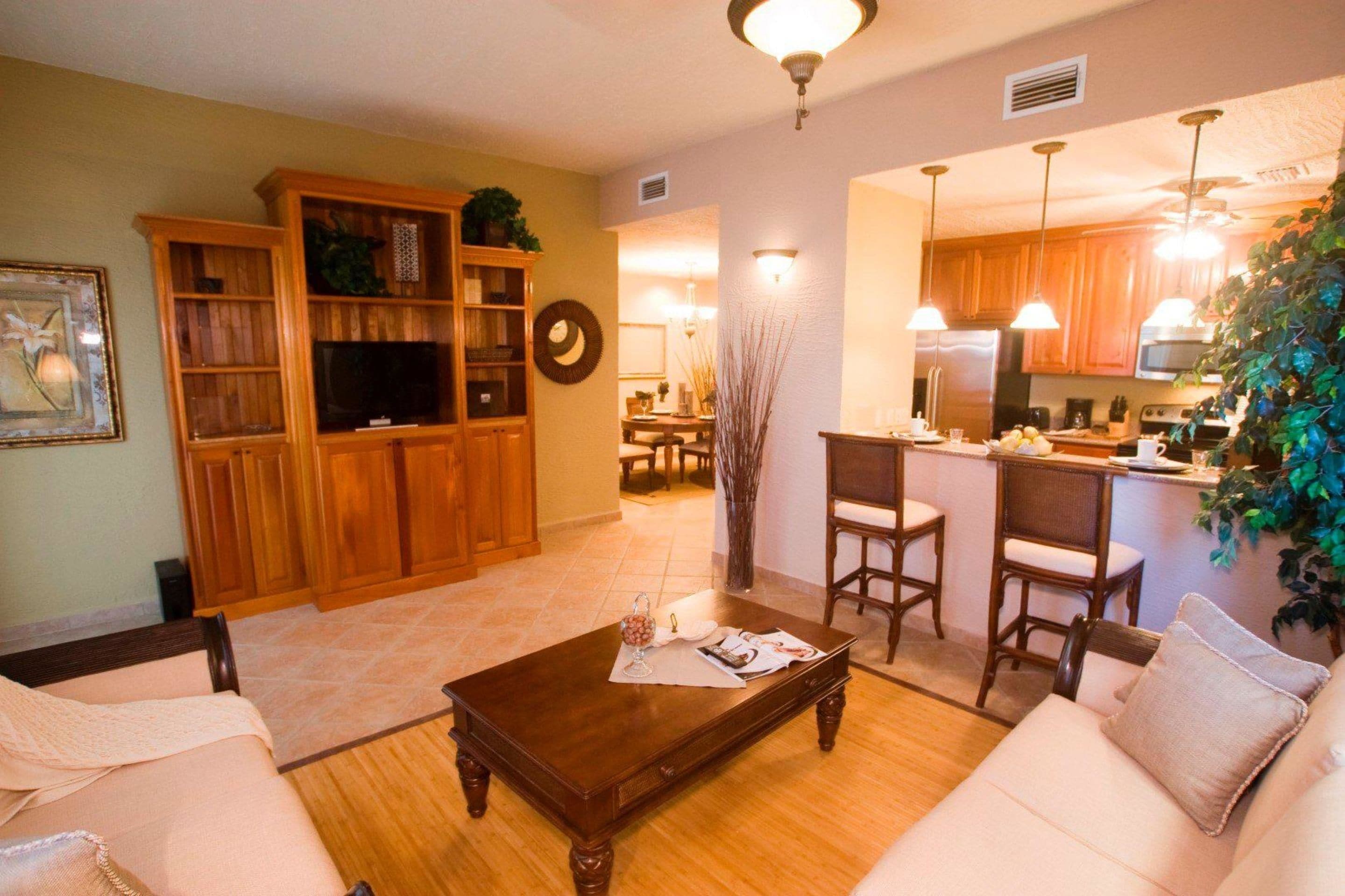 Una sala de estar con una mesa de centro de madera, un sofá y un televisor. Hay una zona de cocina con armarios de madera y un refrigerador.