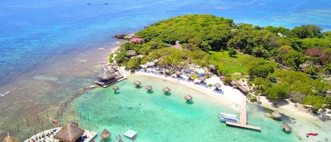 An aerial view of a tropical beach resort with clear turquoise waters, a sandy beach, and lush greenery.