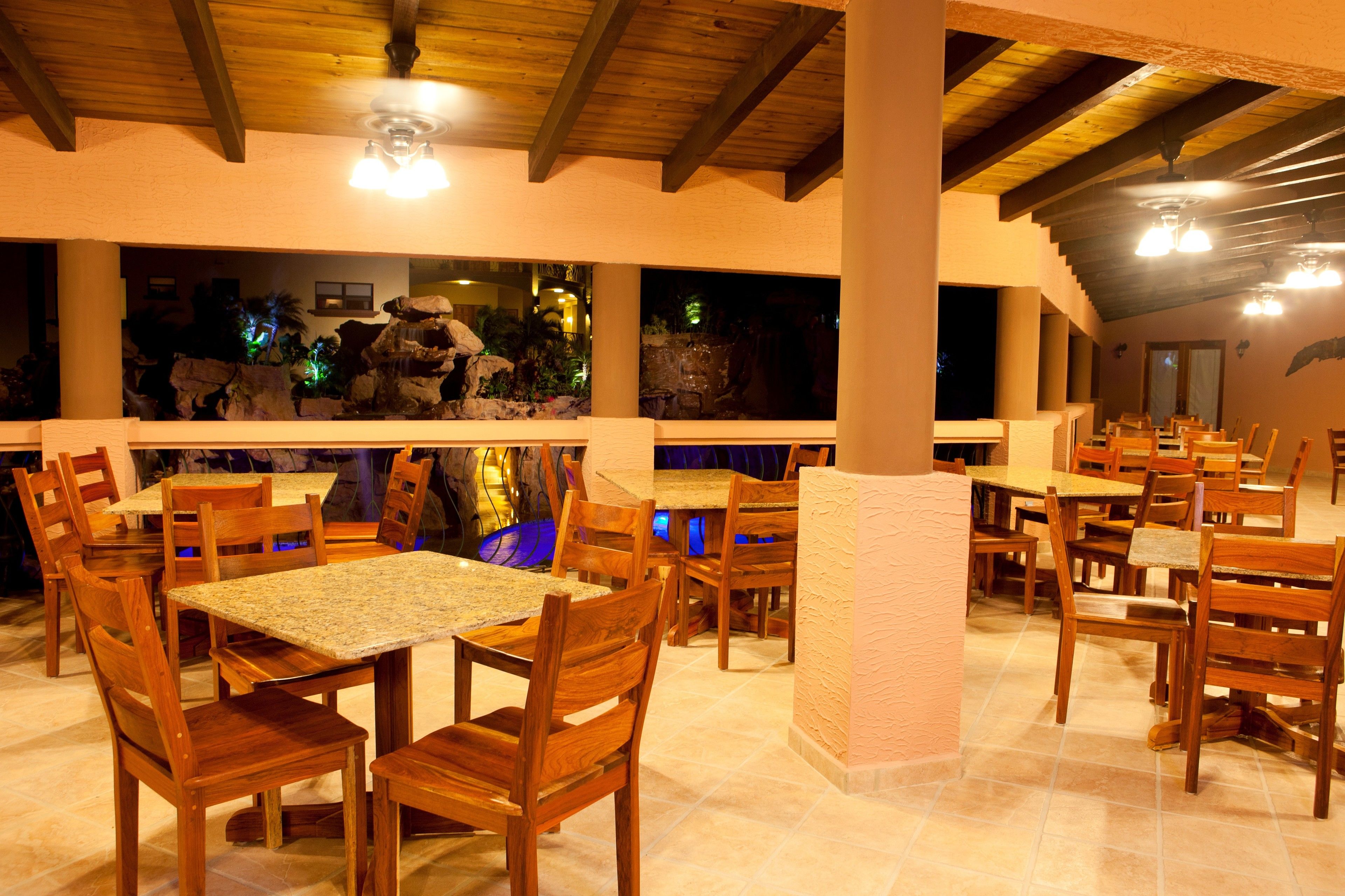 A restaurant with wooden tables and chairs, a stone wall with a rock garden, and a ceiling with wooden beams and lights.