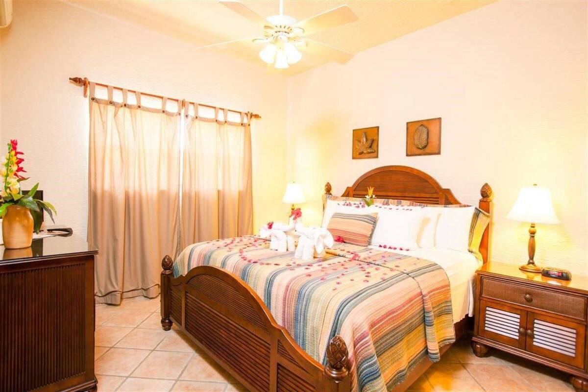 A hotel room with a wooden bed, two bedside tables with lamps, a ceiling fan, and a window with curtains.