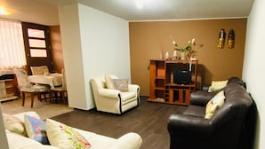 Una sala de estar con sofá, sillón y televisión sobre un soporte de madera. En el fondo, hay una zona de comedor con mesa y sillas.
