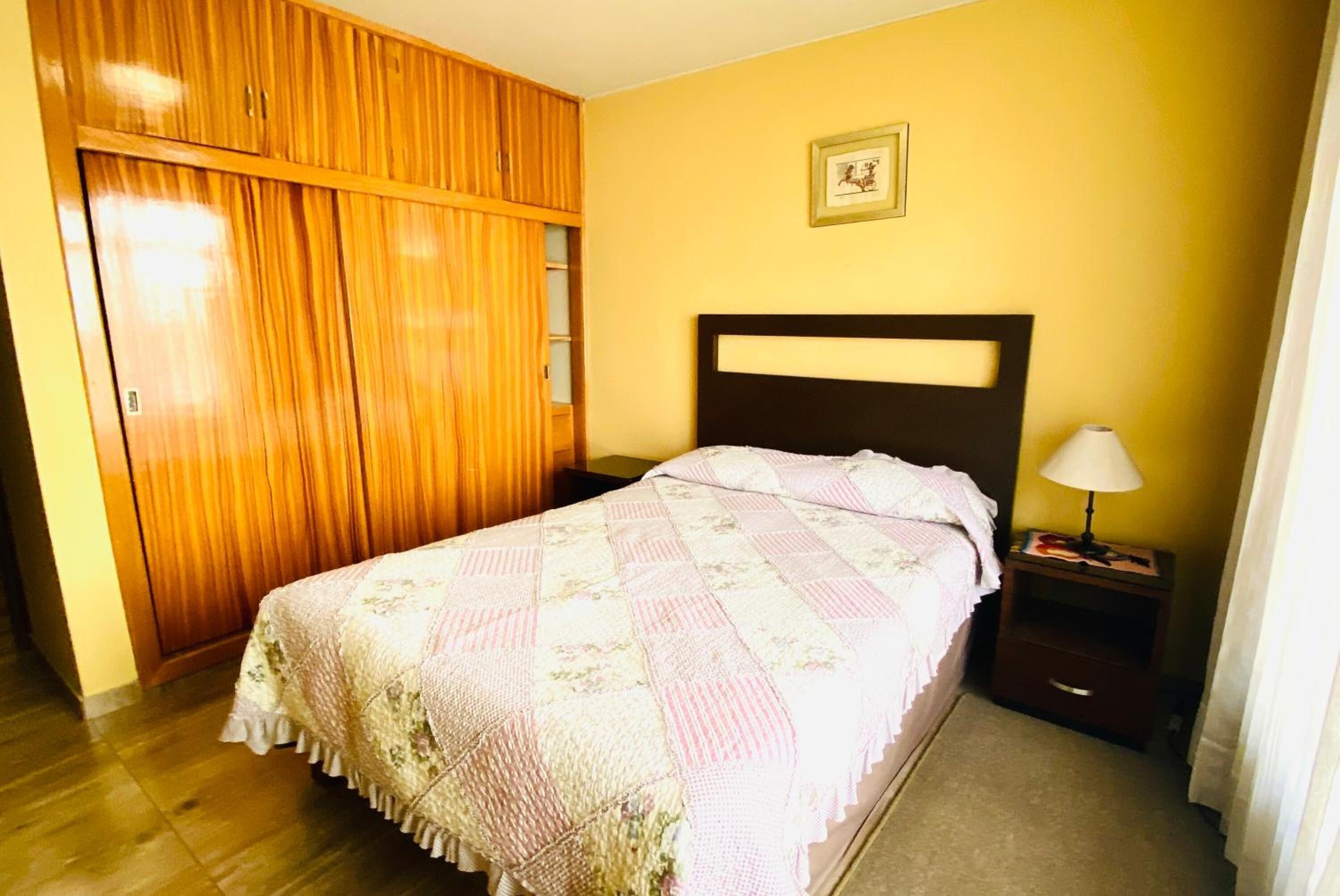 A bedroom with a bed, wooden wardrobe, bedside table, lamp, and a framed picture on the wall.