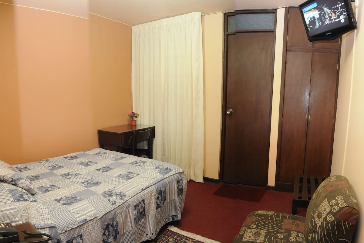 Quarto com cama, escrivaninha, cadeira, televisão e guarda-roupa.