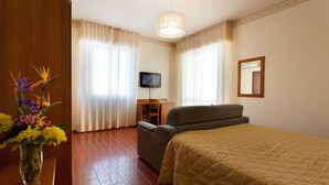 Una habitación de hotel con cama, escritorio, silla, televisor y un jarrón con flores.