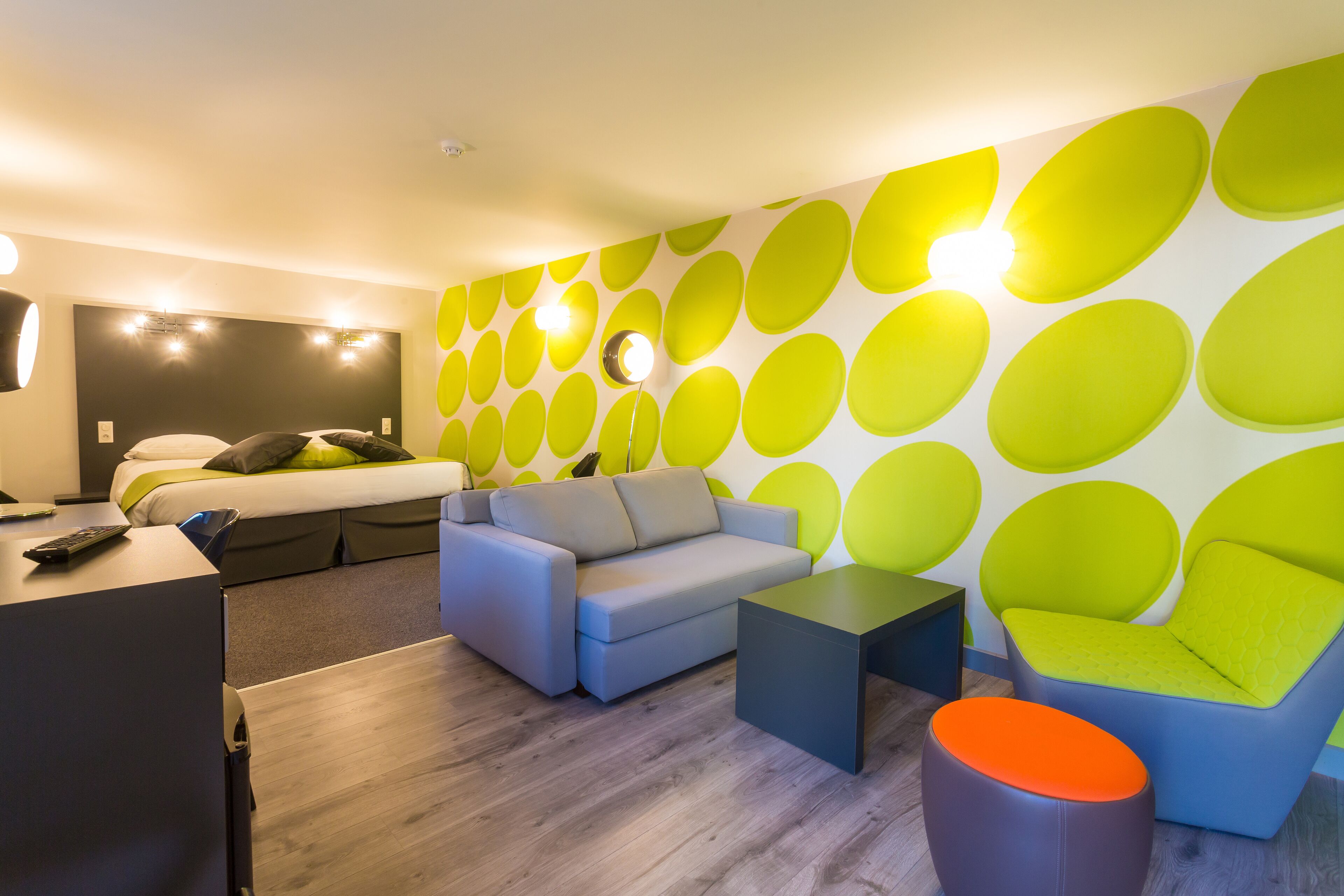 Une chambre d’hôtel moderne avec un canapé gris, un fauteuil bleu, une petite table et un mur vert vif orné de motifs circulaires.