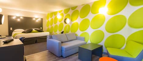A modern hotel room with a grey sofa, a blue armchair, a small table, and a bright green wall with circular patterns.