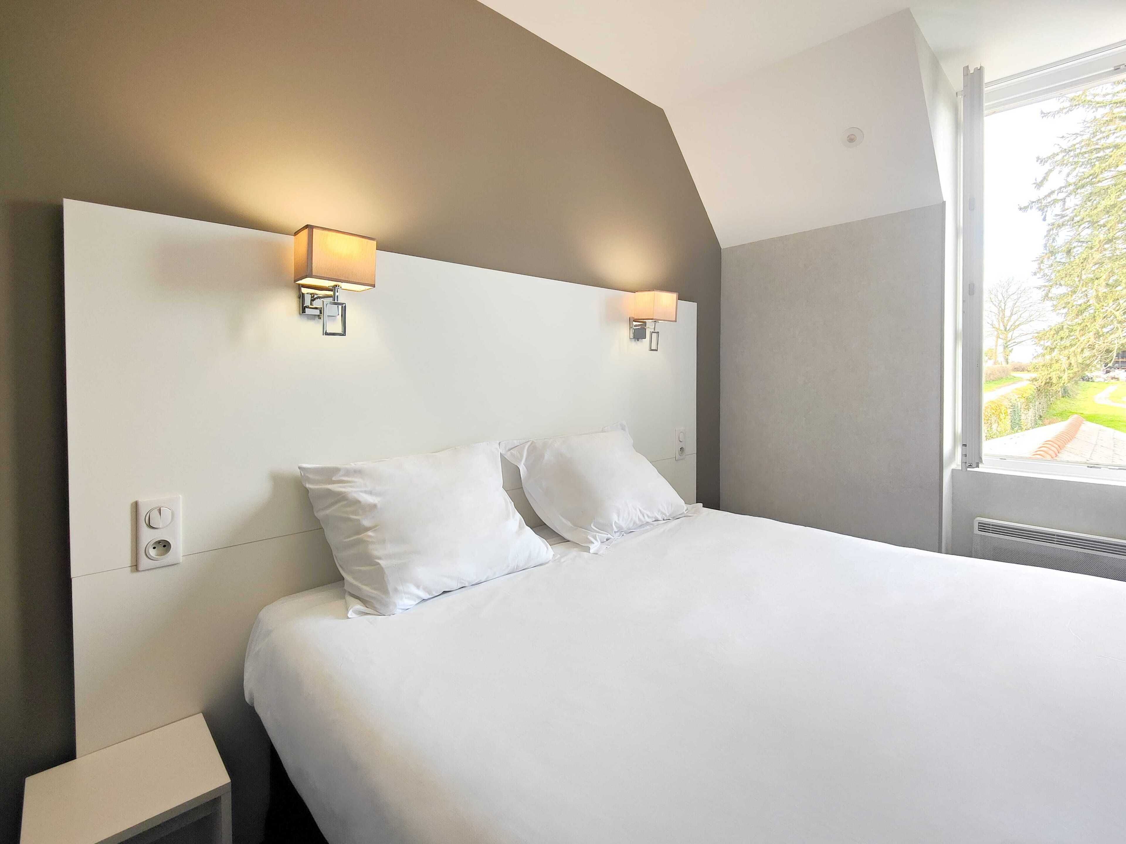 Habitación de hotel con una cama grande, dos lámparas fijadas a la pared, un ventanal con vistas a zonas verdes y un enchufe en la pared.