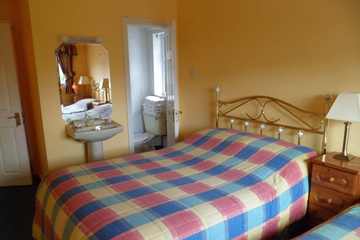 A bedroom with a bed, a bedside table, a lamp, a mirror, a sink, and a door leading to a bathroom.