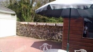 A patio with a stone wall, a wooden cabin, and a table with an umbrella. - The Branches (Galway)