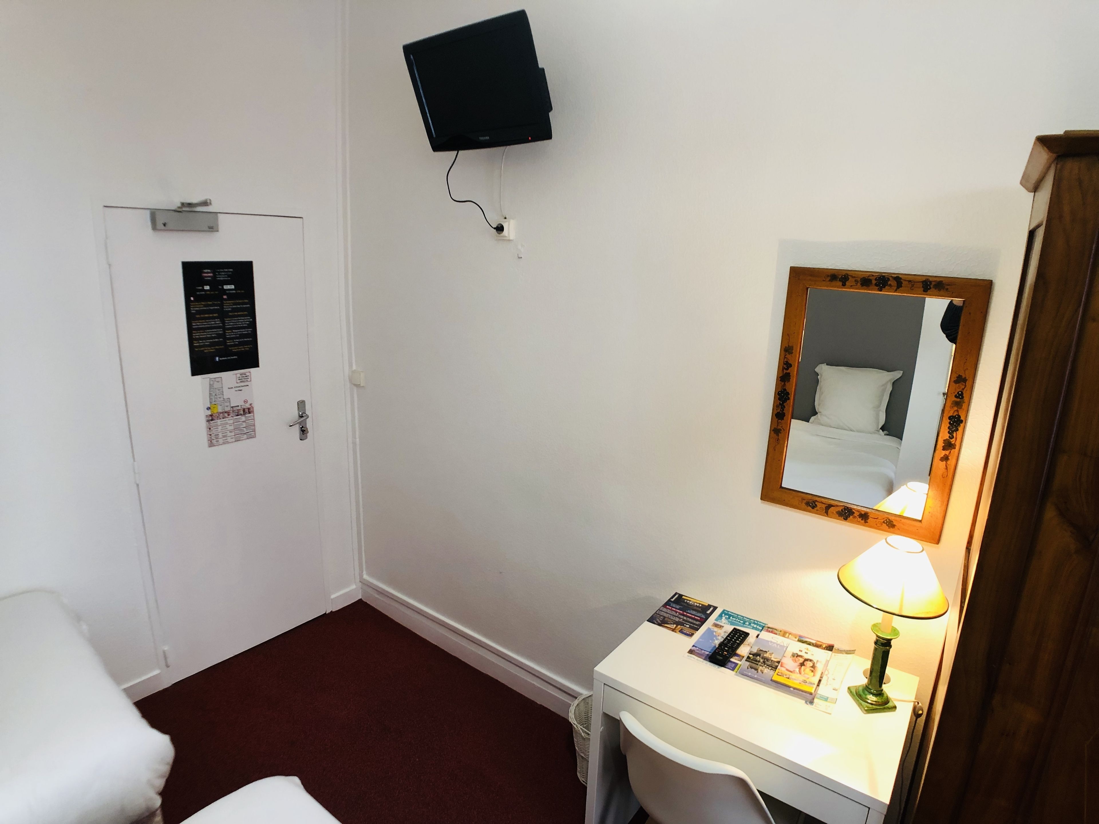 A hotel room with a bed, a desk with a lamp, a mirror, a TV mounted on the wall, and a door with a bulletin board.