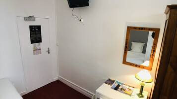 A hotel room with a bed, a desk with a lamp, a mirror, a TV mounted on the wall, and a door with a bulletin board.