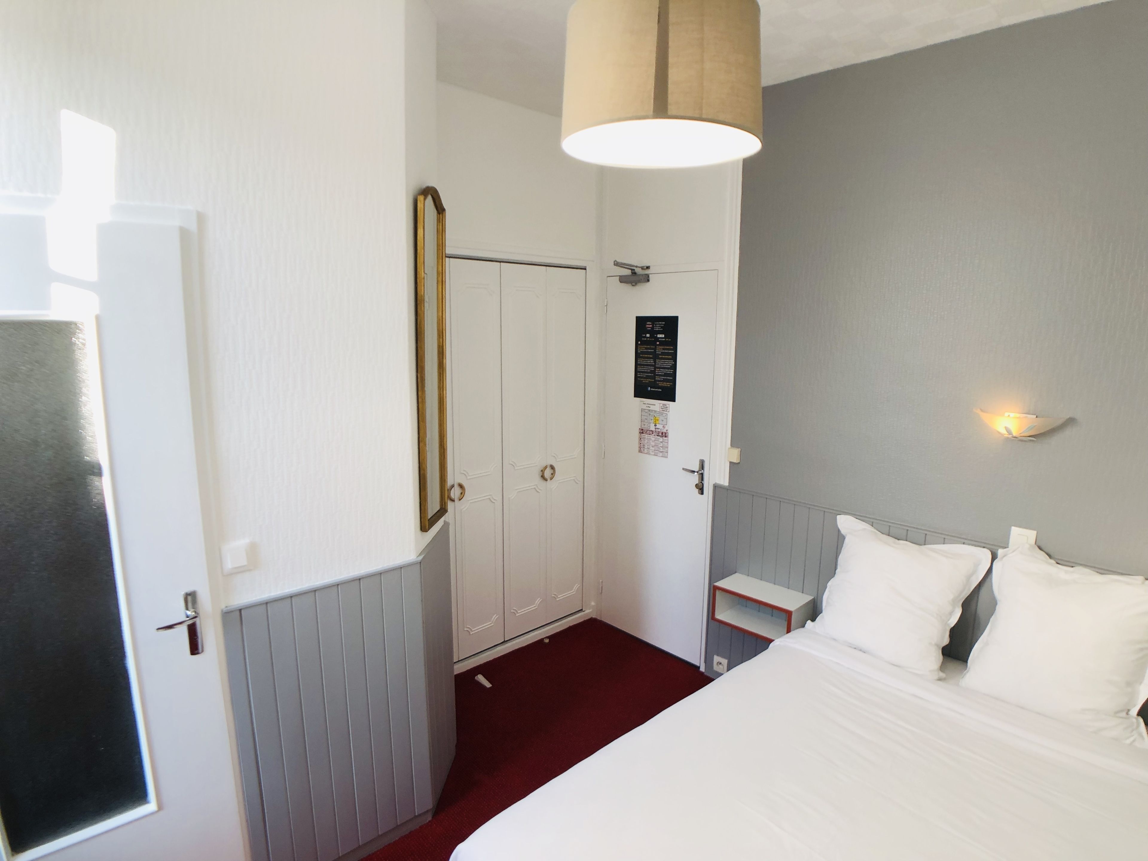 Una habitación de hotel con cama, mesita de noche, espejo, puerta y lámpara fijada a la pared.