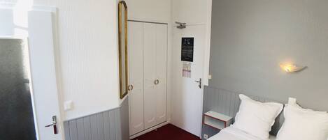 Una habitación de hotel con cama, mesita de noche, espejo, puerta y lámpara fijada a la pared.