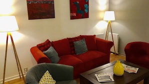Una sala de estar con un sofá rojo, un sillón gris y una mesa de centro con periódicos y un jarrón amarillo. Hay dos lámparas de pie y cuadros abstractos en la pared.