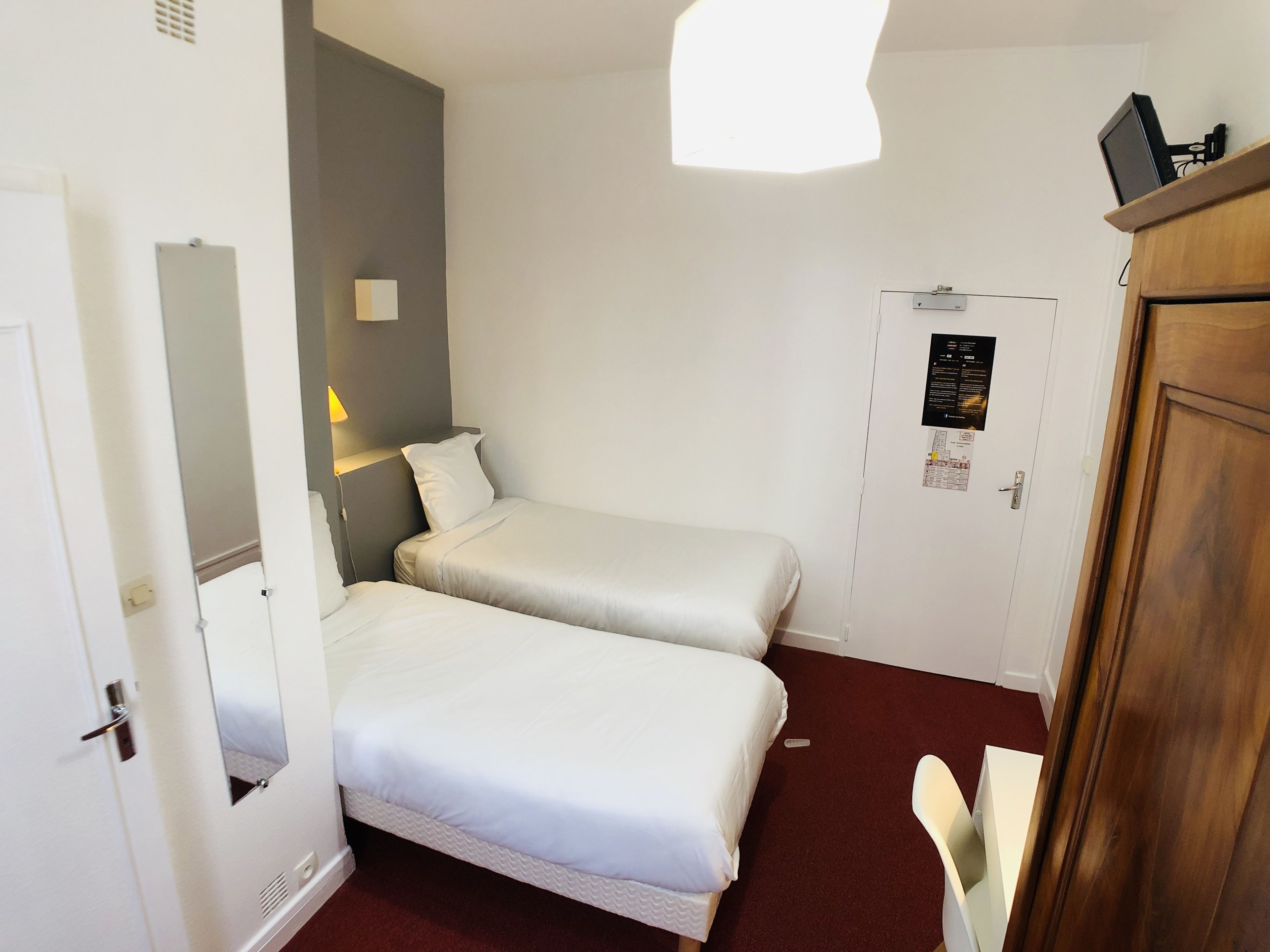 A hotel room with two single beds, a desk, a chair, a wardrobe, and a door with a poster.