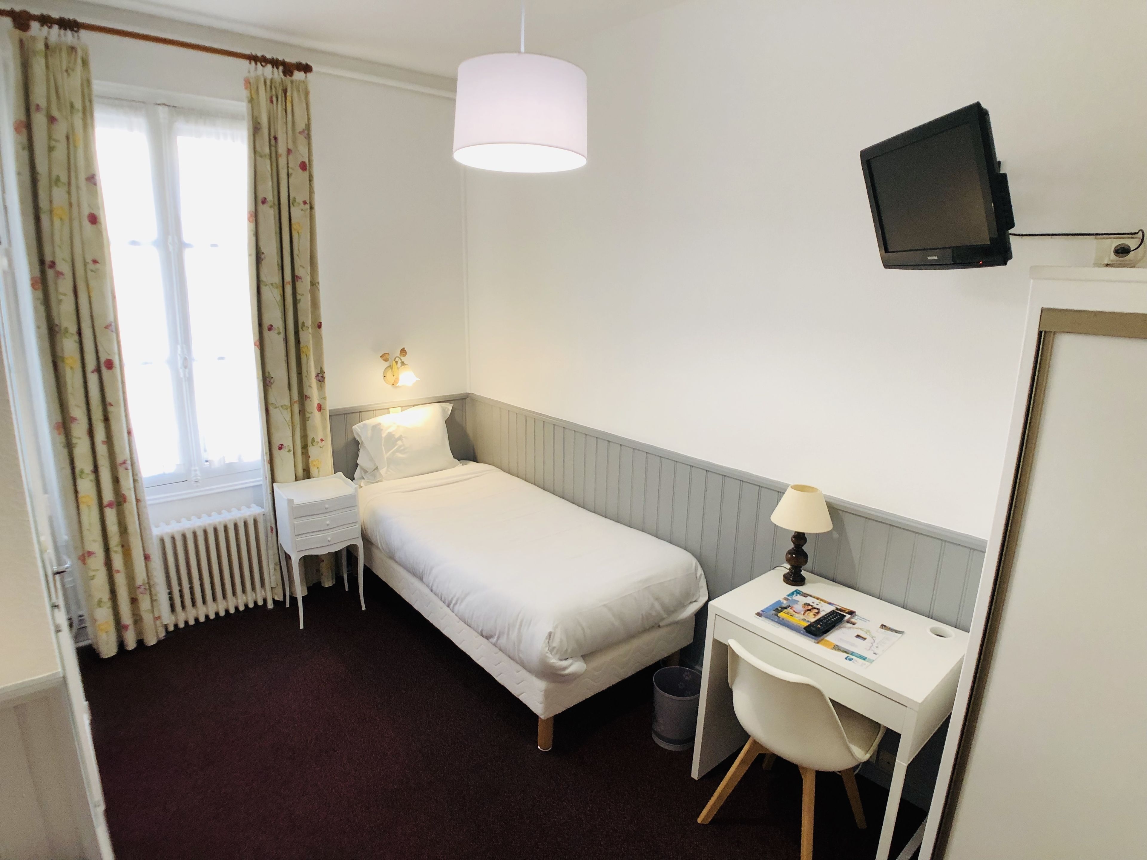 A small, neatly arranged hotel room with a single bed, a desk with a chair, a television mounted on the wall, and a window with curtains.