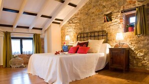 A bedroom with a stone wall, a bed with white bedding and red pillows, a nightstand with a lamp, and a window with curtains. - Posada Santa Ana (Suances)