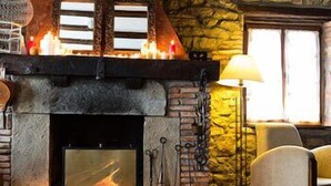 A stone fireplace with a fire, a lamp, and two chairs in a room with a stone wall and a window. - Posada Santa Ana (Suances)