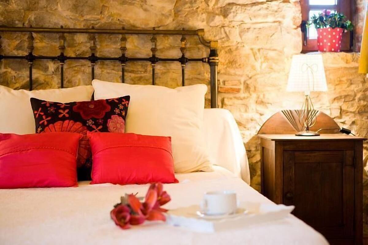 A bed with white linens and red pillows, a bedside table with a lamp, and a stone wall in the background.