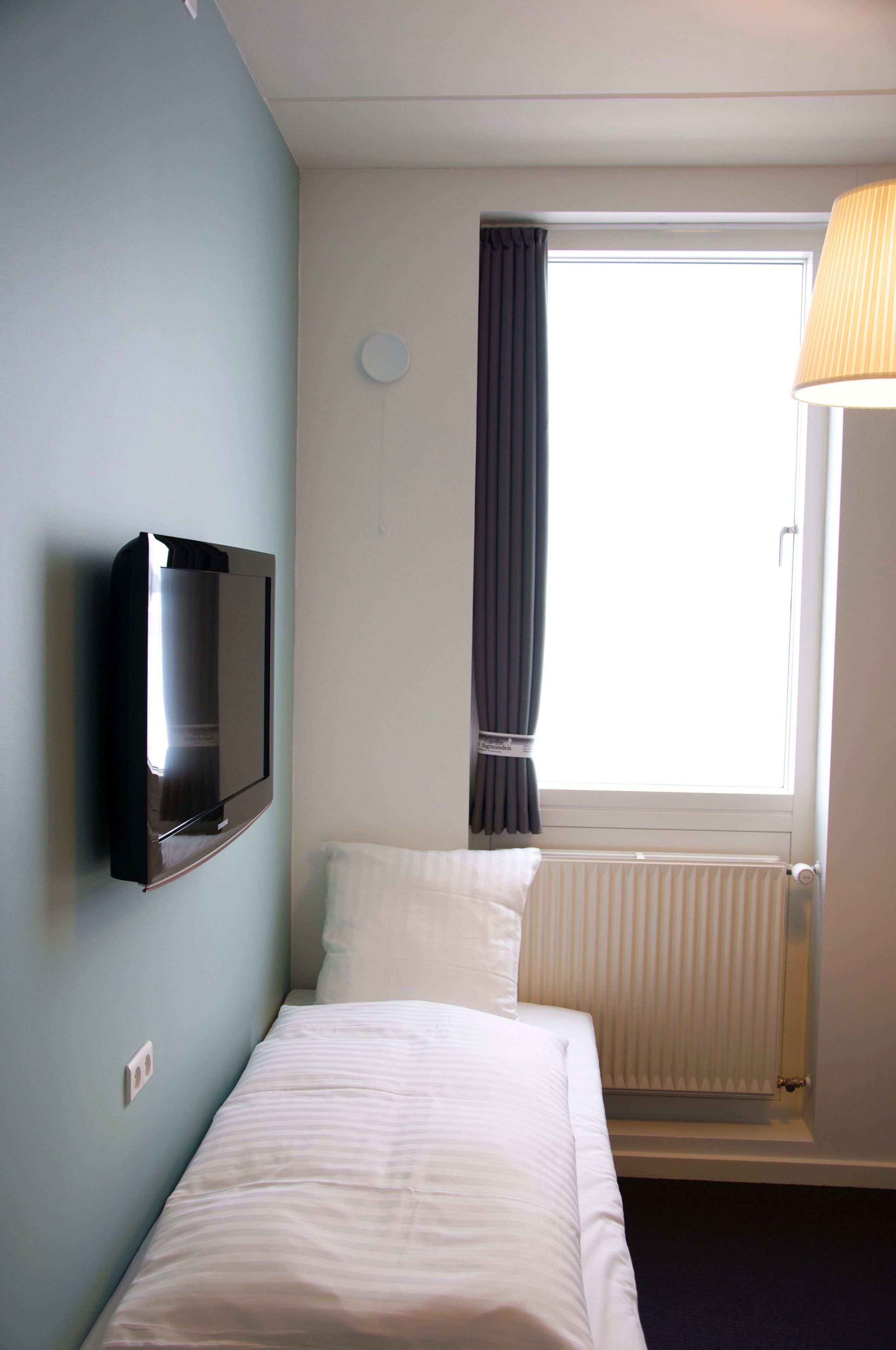 A hotel room with a bed, a flat-screen TV mounted on the wall, a window with curtains, and a radiator.