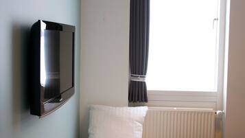 A hotel room with a bed, a flat-screen TV mounted on the wall, a window with curtains, and a radiator.