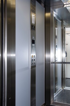 An elevator interior with stainless steel doors, a control panel with buttons, and a glass door. - Motel X (Randers)