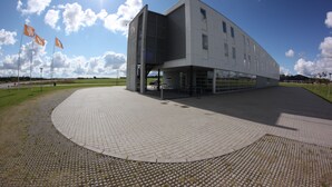 A modern building with a curved facade, large windows, and a paved area in front. - Motel X (Randers)