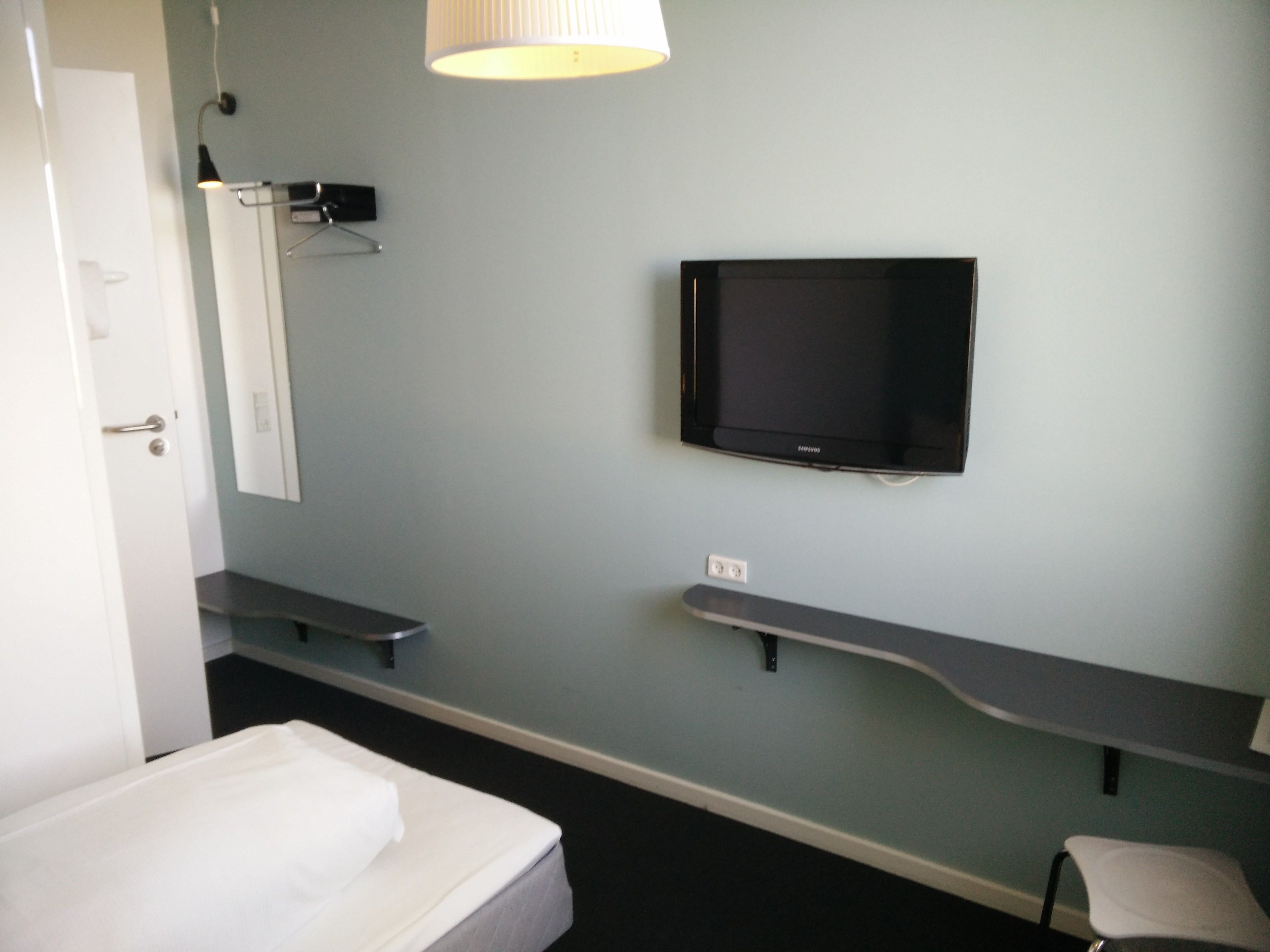 A hotel room with a bed, a TV mounted on the wall, a bedside table, and a lamp.