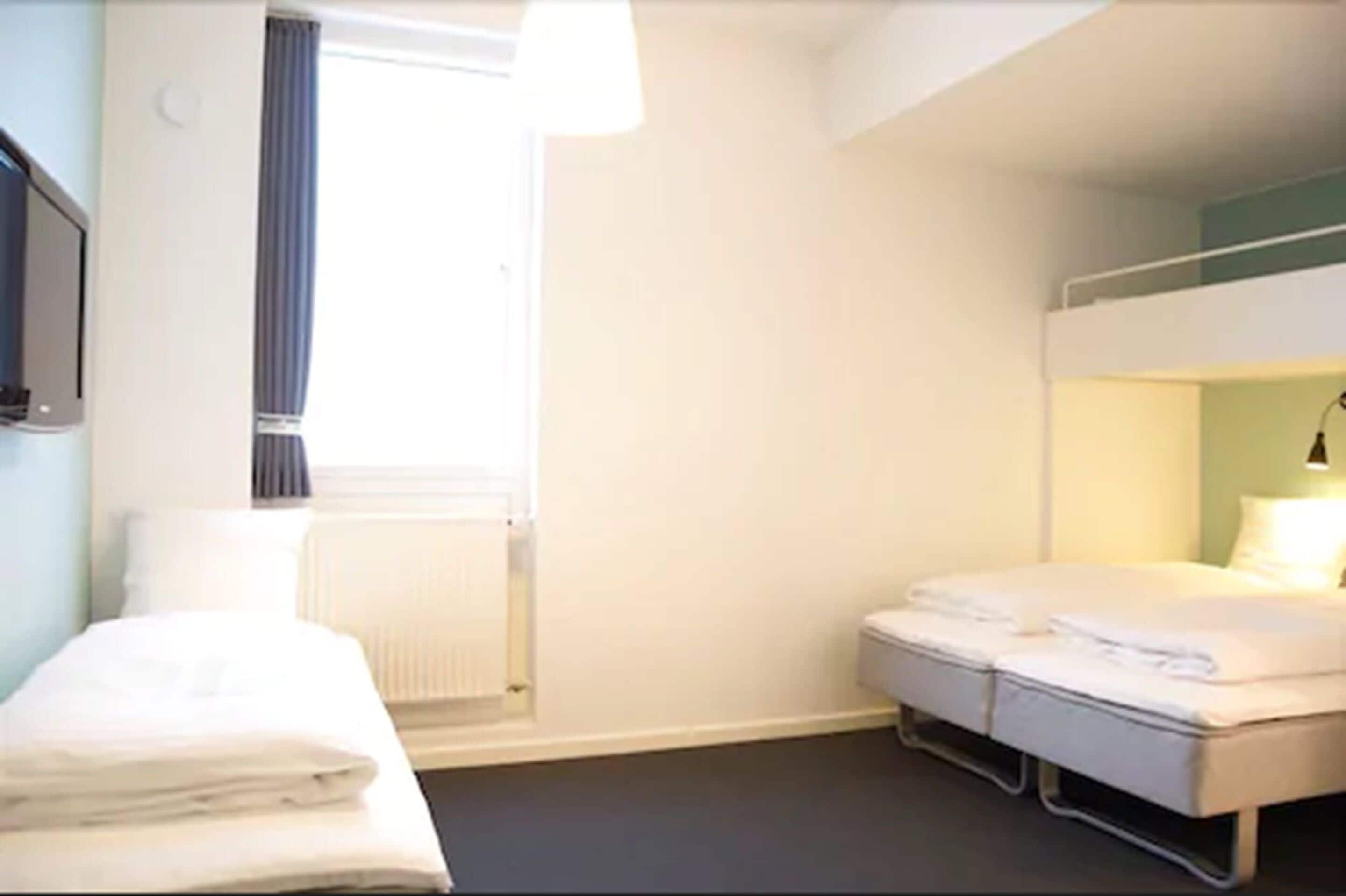 A hotel room with a bunk bed, a bedside table, a radiator, and a window with curtains.