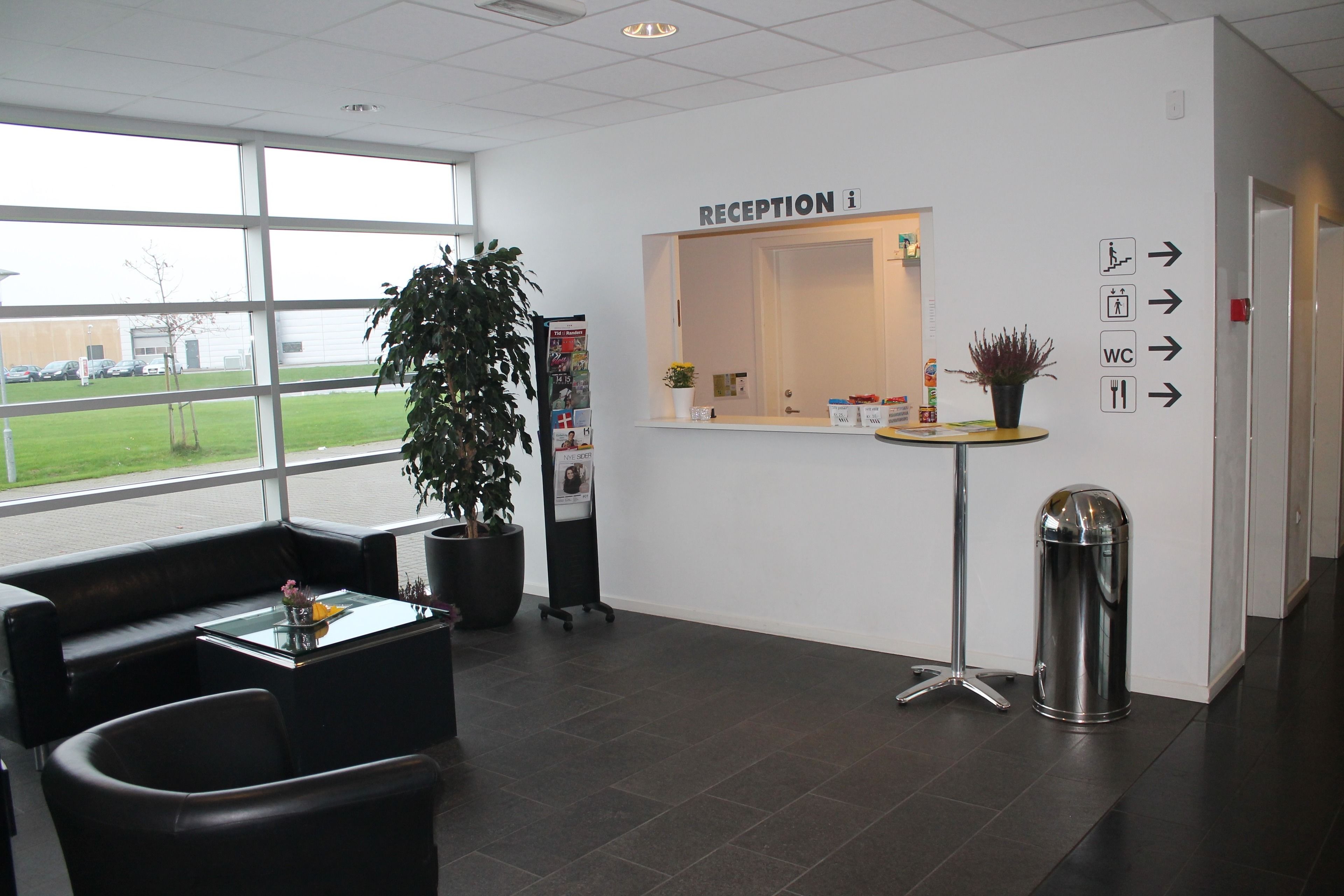 A reception area with a reception desk, directional signs, a trash can, a potted plant, and a seating area with a glass-top table and chairs.