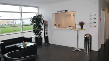 Un espace d’accueil comprenant un comptoir de réception, des panneaux d’orientation, une poubelle, une plante en pot et un coin salon avec une table et des chaises à plateau en verre.