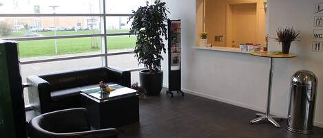 A reception area with a reception desk, a large window with a view of the parking lot, a potted plant, and a trash bin.
