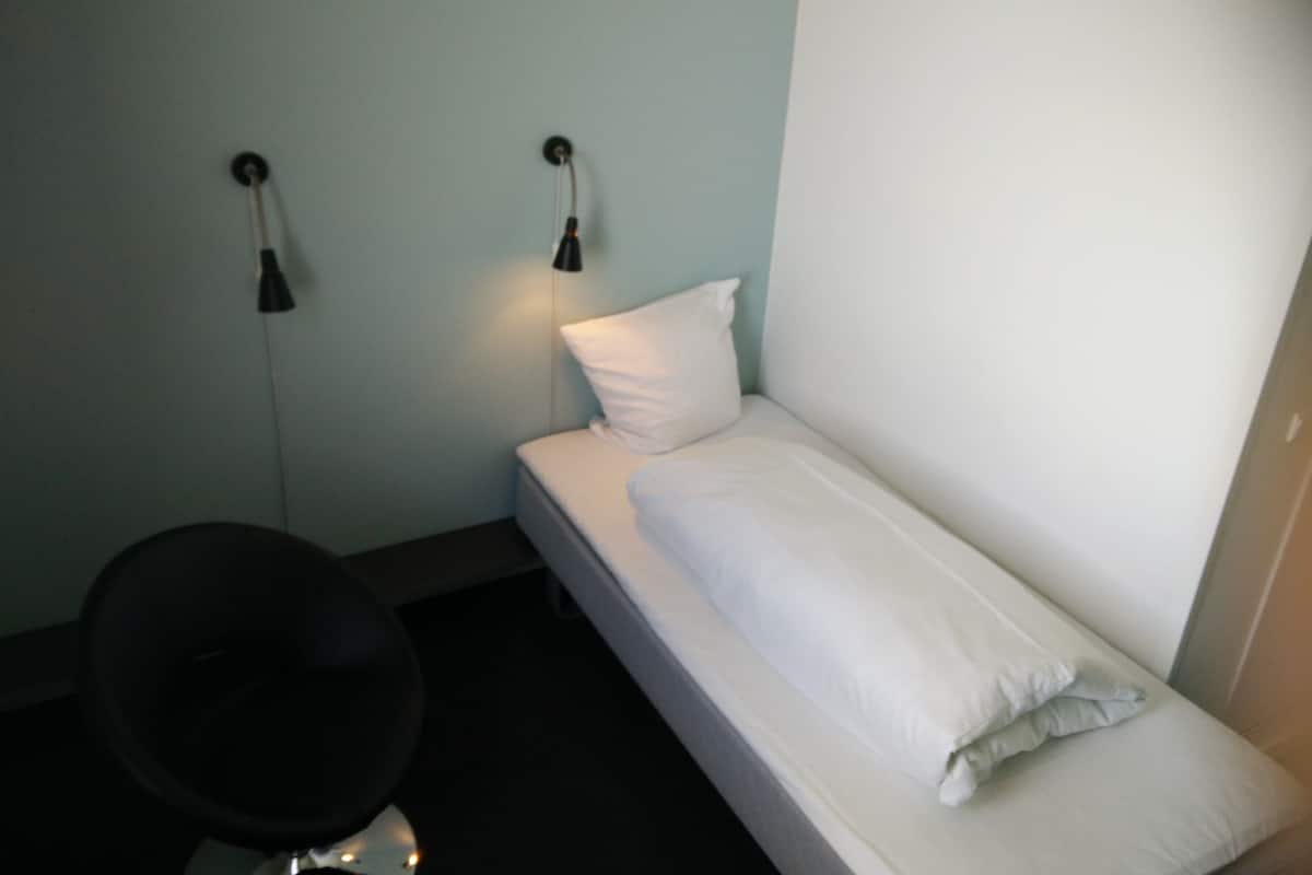 Una habitación pequeña con cama individual, una lámpara sencilla sobre la cama y una silla en la esquina.