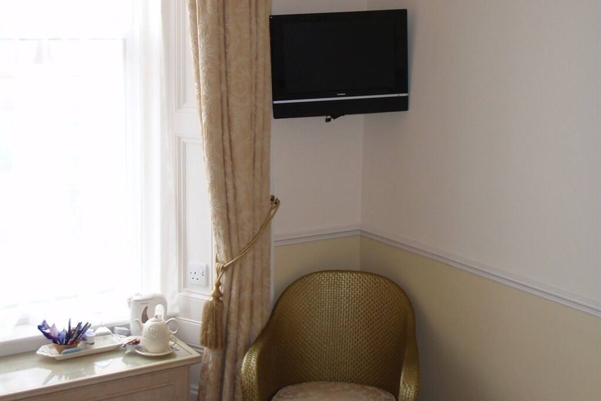 A room with a television mounted on the wall, a wicker chair, a window with curtains, and a dresser with a tea set.