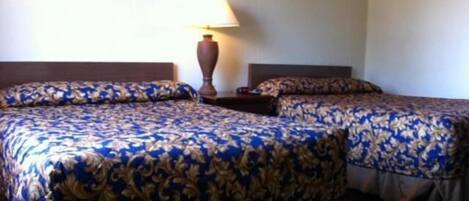 A hotel room with two beds, each with a patterned bedspread, a nightstand with a lamp, and a plain wall.