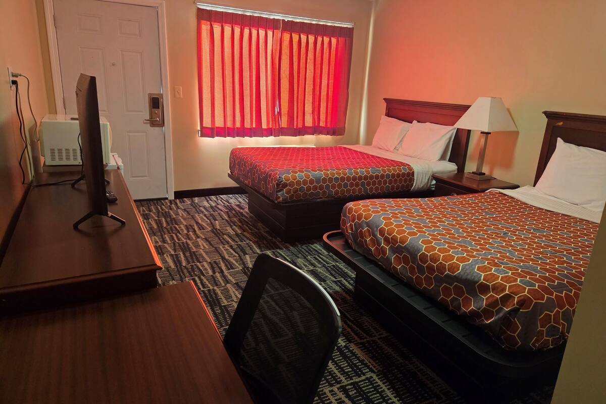 A hotel room with two beds, a desk with a computer, a lamp, and a window with red curtains.